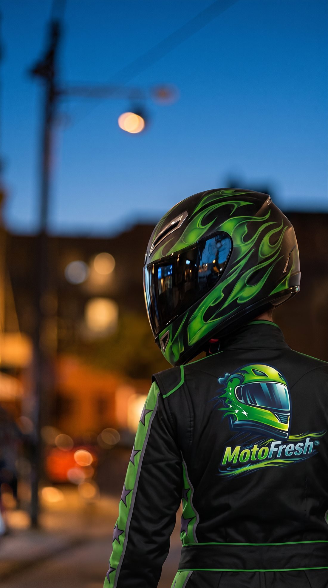 Motociclista de espaldas con casco de llamas verdes y chamarra MotoFresh en una calle de noche.